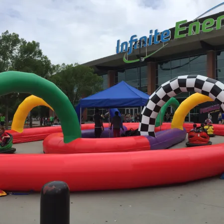 Outdoor inflatable go-kart track with colorful arches and riders in front of Infinite Energy building on cloudy day
