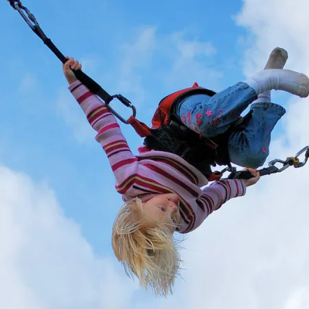 Child in striped sweater and jeans swinging upside down on a harness against a cloudy blue sky.