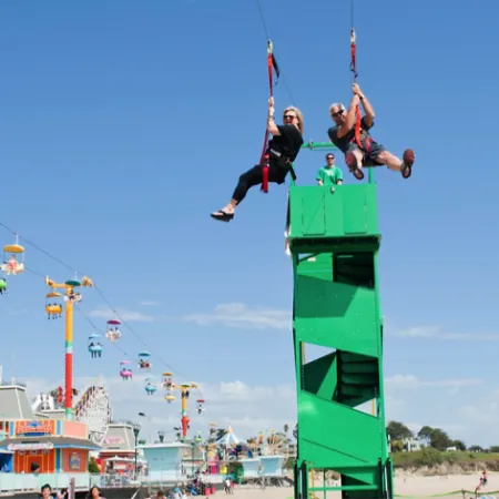 Two people ziplining from a green tower over a lively amusement park with sky chairs and rides on a sunny day
