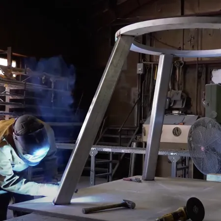 Welder wearing protective gear welding a large metal frame in an industrial workshop with tools on table.