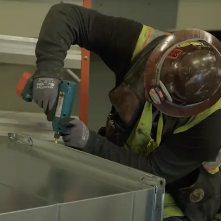 Construction worker wearing safety gear using a power drill on metal ductwork indoors with an orange ladder in background