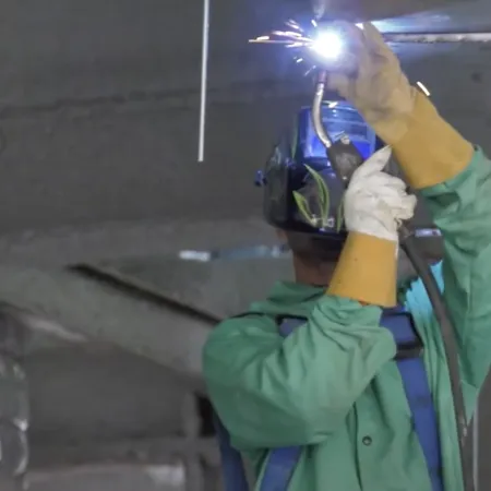 Welder wearing protective gear welding a metal structure indoors with sparks flying from the welding torch.