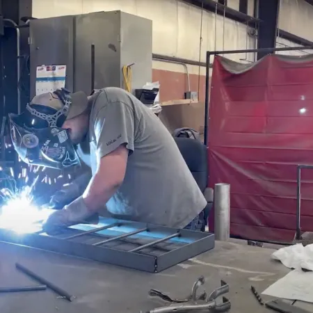 Welder wearing protective gear sparks metal welding in an industrial workshop with tools and equipment.