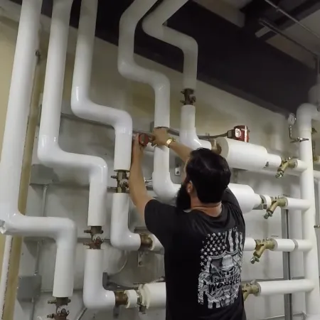 Technician adjusting valves on complex white industrial piping system in a utility room with fluorescent lighting