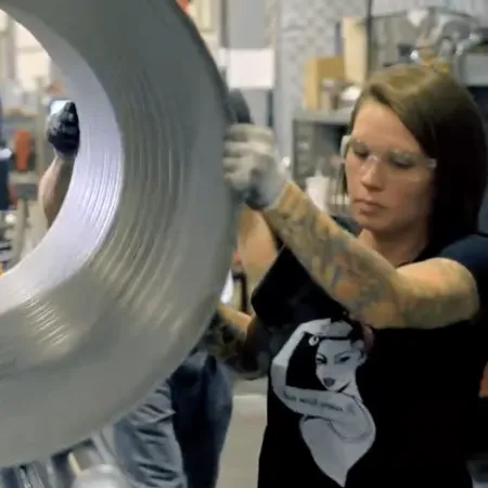 Worker handling a large metal coil in an industrial manufacturing facility wearing safety glasses and gloves.