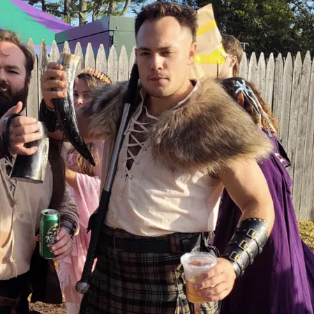 Two men in medieval attire holding drinking horns and cups, standing outdoors near tents and a wooden fence.