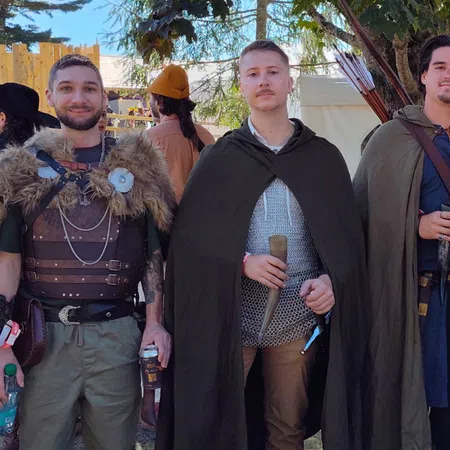 Five men dressed in medieval costumes with cloaks and weapons posing outdoors in daylight under trees.