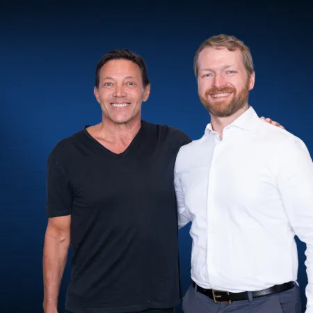 Two men smiling, one in a black t-shirt and the other in a white dress shirt, posing against a blue background