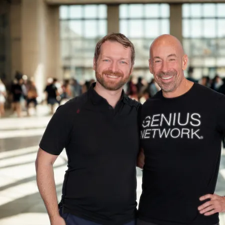 Two smiling men standing close together in a bright indoor space with large windows and people in the background.
