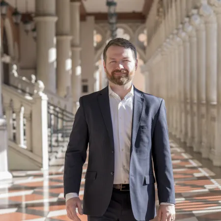 Man in a dark suit walking confidently through a sunlit colonnade with patterned tile flooring.