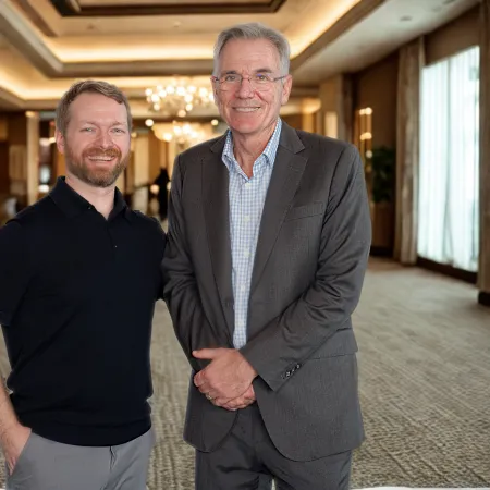 Two men smiling and posing in a well-lit, elegant hallway with chandeliers and large windows.