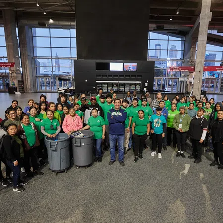 Large diverse group of people posing indoors with recycling bins in a spacious hall with large windows.