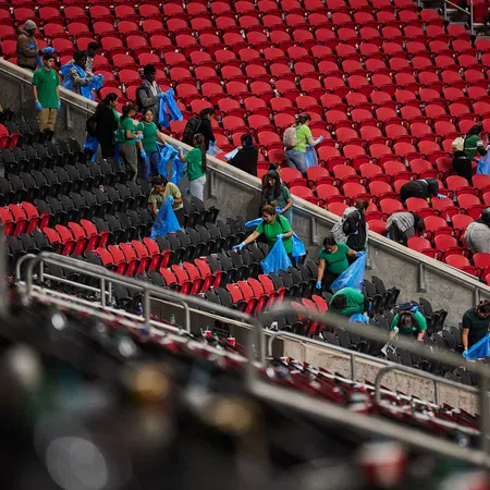 Volunteers in green shirts cleaning empty red and black stadium seats holding blue trash bags.