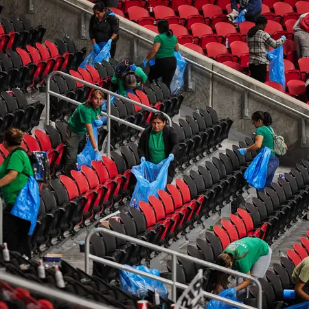 Group of people in green shirts cleaning red and black stadium seats holding blue trash bags and gloves.