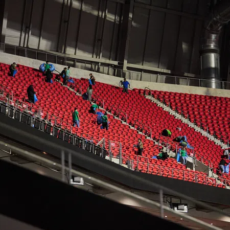 Workers in green and blue uniforms clean red stadium seats in a large indoor sports arena under bright lights.
