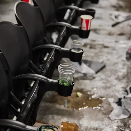 Row of black stadium seats with various empty drink cups and trash scattered on wet concrete floor.