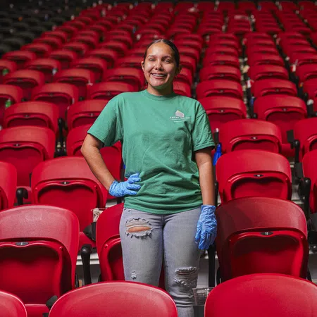 Young woman in green shirt and blue gloves standing amid rows of empty red stadium seats, smiling at camera.