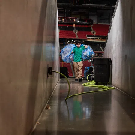 Person carrying large bags of recyclable plastic bottles through a stadium corridor with equipment and hoses present.