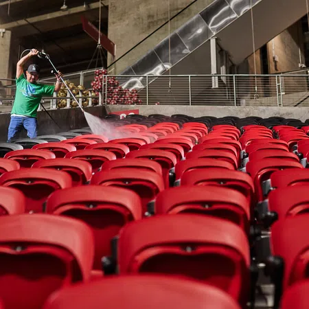 Worker in green shirt pressure washing rows of red stadium seats in a large indoor arena.