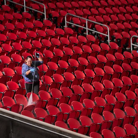 Two workers in blue shirts cleaning rows of red stadium seats wearing gloves and using spray equipment.