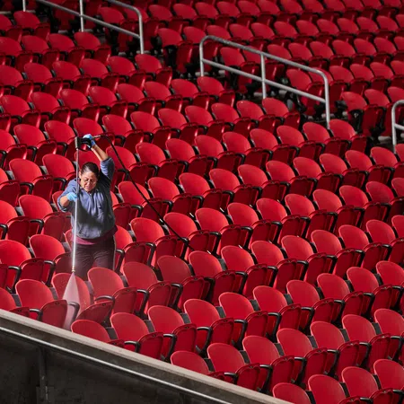 Two workers cleaning rows of empty red seats in a large indoor arena or stadium with spray bottles and cloths.