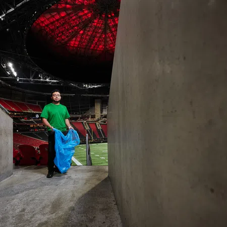 Man in green shirt holds blue trash bag in stadium tunnel with red-lit ceiling and empty seats.