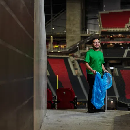 Person in green shirt holding a blue trash bag standing in an empty stadium corridor with red and black seats.