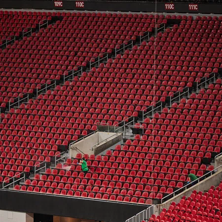 Empty red stadium seats with a few people scattered and stairways dividing sections under daylight.