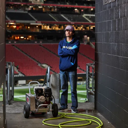 Man in casual clothes standing with arms crossed next to lawn equipment in a stadium tunnel.