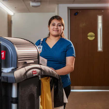 Healthcare worker in blue scrubs pushing a cleaning cart in a hospital hallway with a smile.