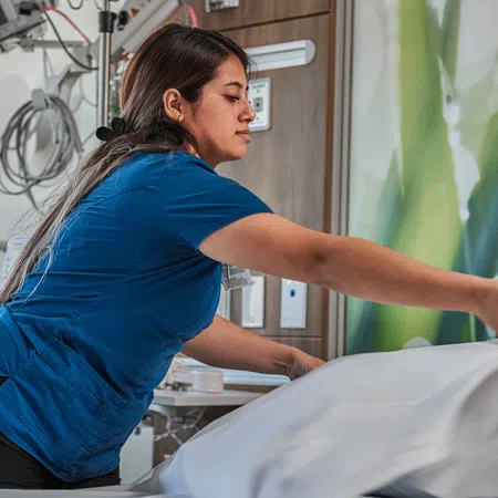 Healthcare worker in blue scrubs making a hospital bed with white linens in a patient room.