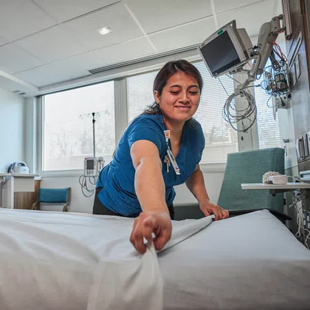 Nurse making a hospital bed in a bright patient room with medical equipment and Piedmont logo on screen.