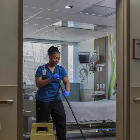 Healthcare worker in blue scrubs mopping hospital room floor with wet floor caution sign in foreground.
