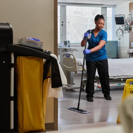 Healthcare worker mopping floor in a clean hospital room with medical equipment and caution sign visible.