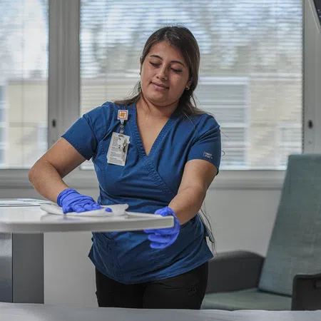 Healthcare worker in blue scrubs cleaning a hospital room table wearing purple gloves during sanitation.