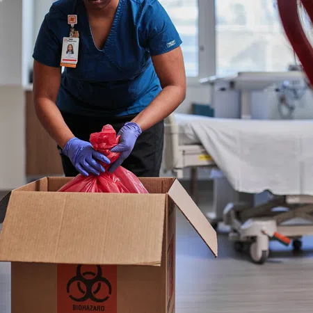 Healthcare worker in blue scrubs placing a red biohazard bag into a cardboard box in a medical facility.