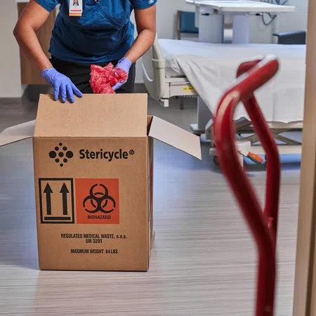 Healthcare worker in gloves disposing red biohazard bags into a Stericycle medical waste box in hospital room.