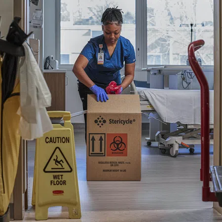 Healthcare worker wearing gloves disposing medical waste into a Stericycle biohazard box in hospital room.