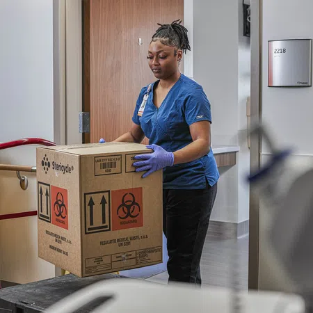 Healthcare worker in scrubs handling a biohazard waste box inside a medical facility hallway.