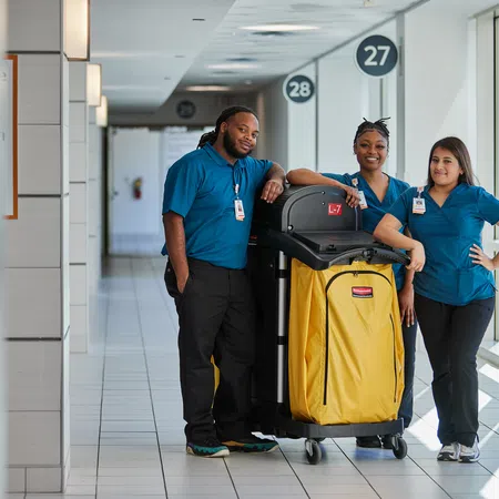 Three janitors in blue uniforms standing with a yellow cleaning cart in a bright hallway with numbered signs.