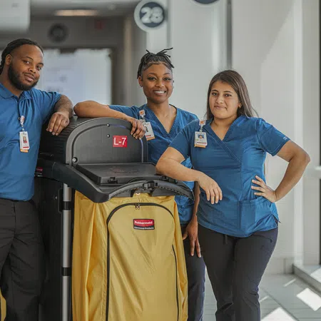 Three janitorial staff in blue uniforms pose with a yellow cleaning cart in a bright hallway.