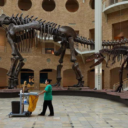 Museum hall with large dinosaur skeletons and a janitor pushing a cleaning cart on tiled floor under high ceilings.