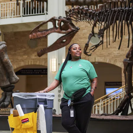 Museum staff member in green shirt standing beside cleaning cart near dinosaur skeleton exhibits indoors.