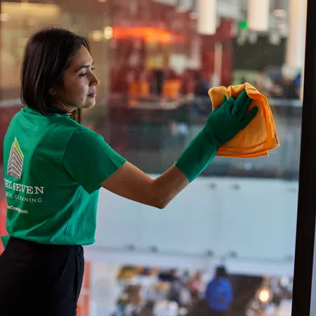 Woman wearing green gloves and shirt cleaning a large glass window with an orange cloth indoors.