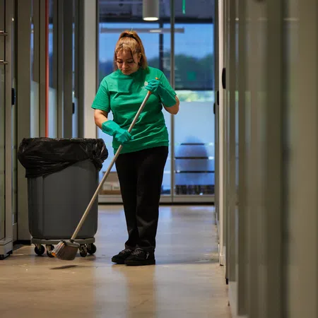 Woman in green shirt mopping floor in a brightly lit modern office hallway with a trash bin nearby