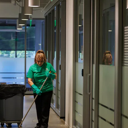Janitor wearing green cleaning a hallway floor with mop and gloves beside trash bin in modern office space.