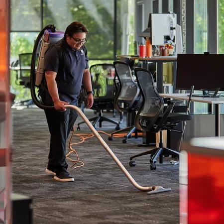 Janitor vacuuming carpeted floor in modern office with large windows and ergonomic chairs during daytime.