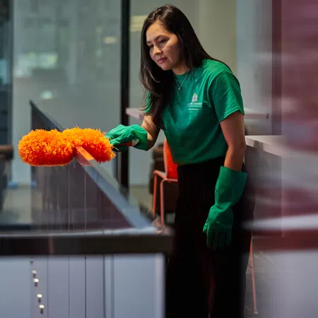 Woman wearing green gloves dusting a glass railing with an orange duster in a modern indoor space.