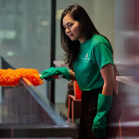 Woman in green shirt and gloves dusting a glass surface with an orange duster in an indoor setting