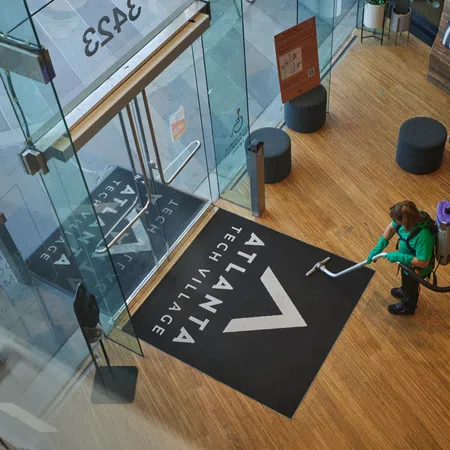 Person vacuuming wooden floor near glass entrance with Atlanta Tech Village logo mat inside building lobby